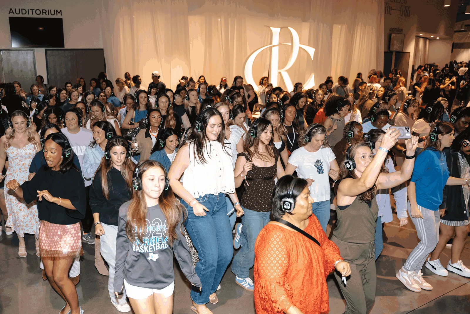 A packed hall of women dancing in silent disco headphones in front of a glowing stage logo