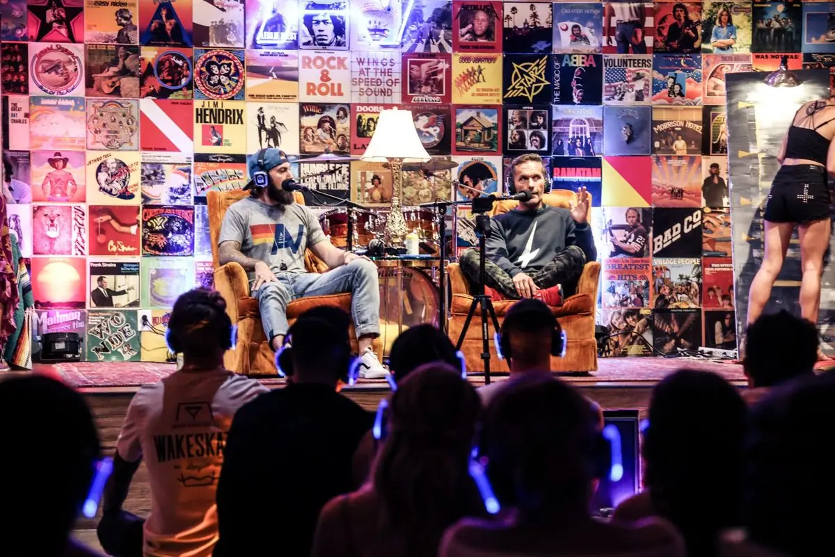 Hosts on stage in front of a record-poster wall, audience listening in headphones