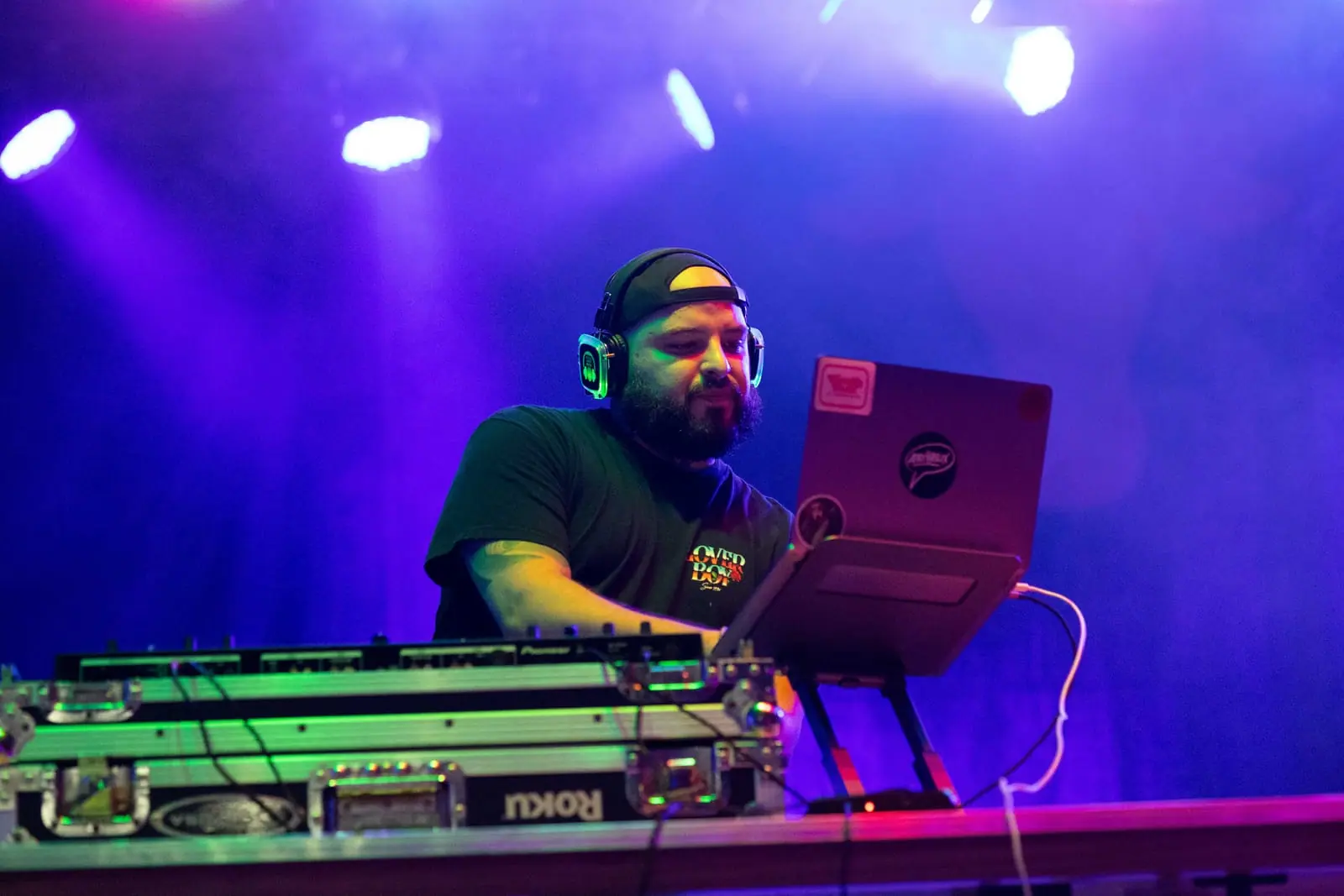 DJ in a ball cap working the laptop and controller under purple light