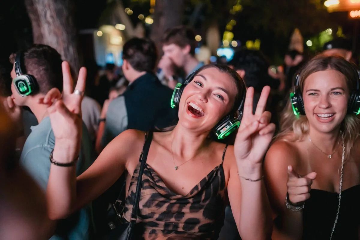 Two friends mid-laugh, peace signs up, headphones glowing