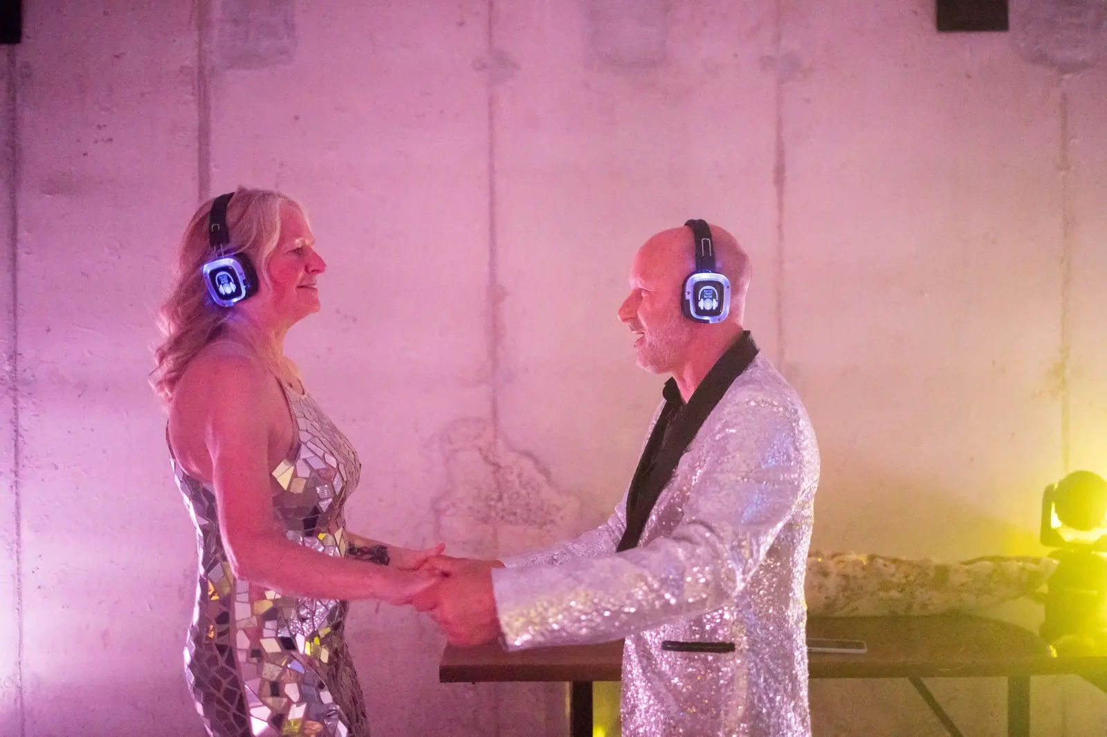 A couple in sparkling outfits, headphones on, in a pink-lit corner of the reception