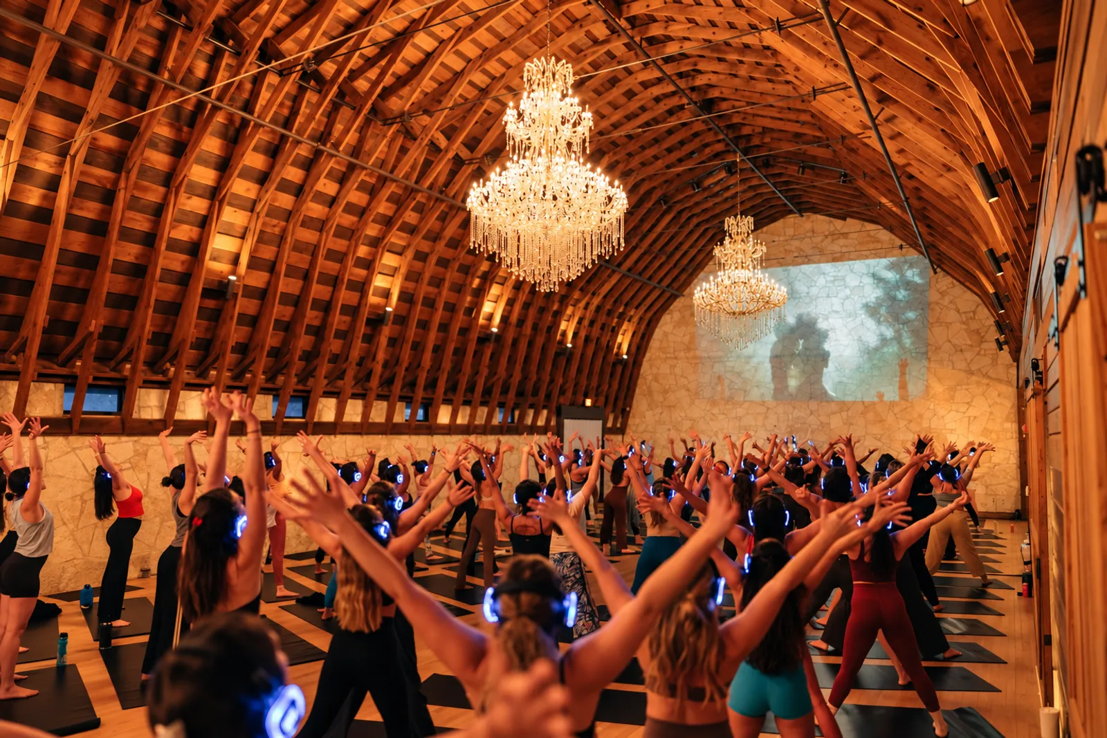 A packed barn yoga class, arms raised under crystal chandeliers, blue headphones glowing