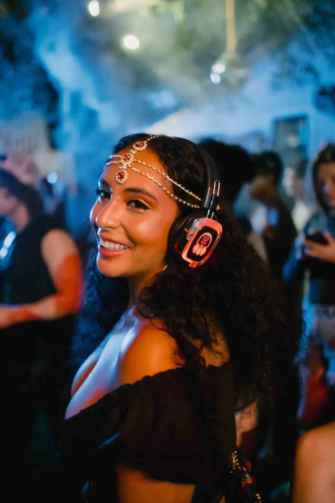A smiling guest in a jeweled headdress, headphones glowing, haze behind